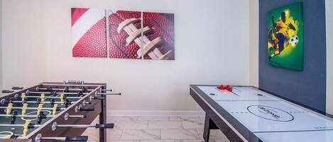 A room with a foosball table and shuffleboard table, both in neutral colors, with a simple tiled floor and plain walls.