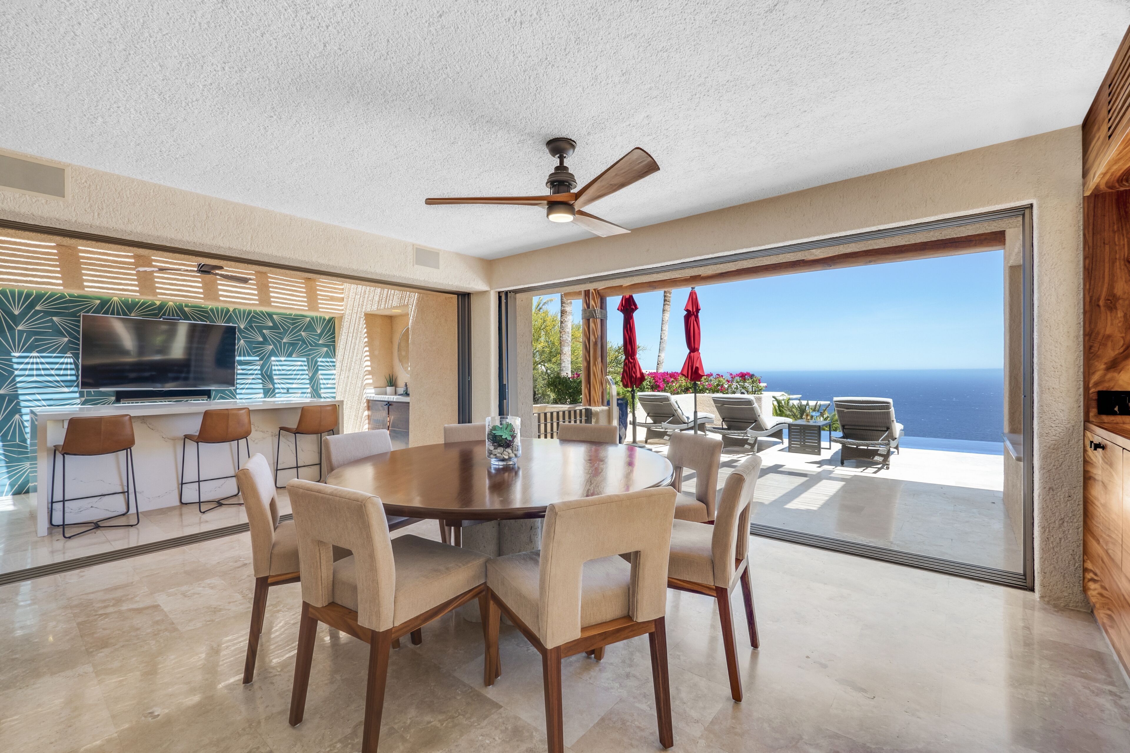 A modern kitchen with a large TV, a dining area with a round table and chairs, and a view of the ocean through large sliding glass doors.