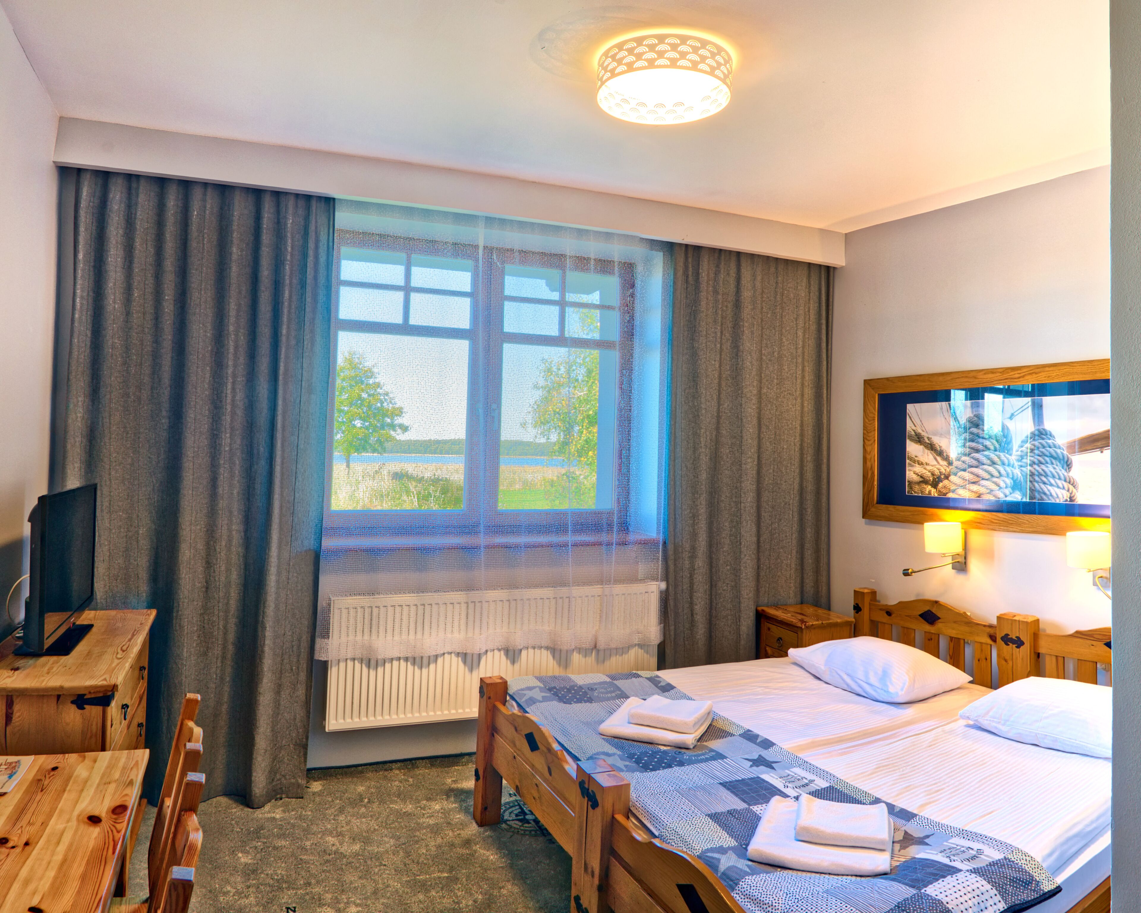 Quarto de hotel com cama, cabeceira de madeira, escrivaninha com cadeira, televisão, janela com cortinas e um quadro na parede.