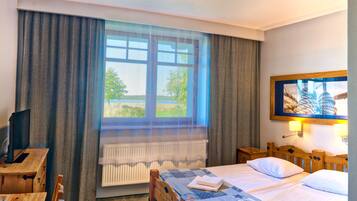 Quarto de hotel com cama, cabeceira de madeira, escrivaninha com cadeira, televisão, janela com cortinas e um quadro na parede.