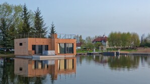 Una cabaña de madera moderna junto a un lago, con su reflejo, rodeada de árboles y una casa a lo lejos.