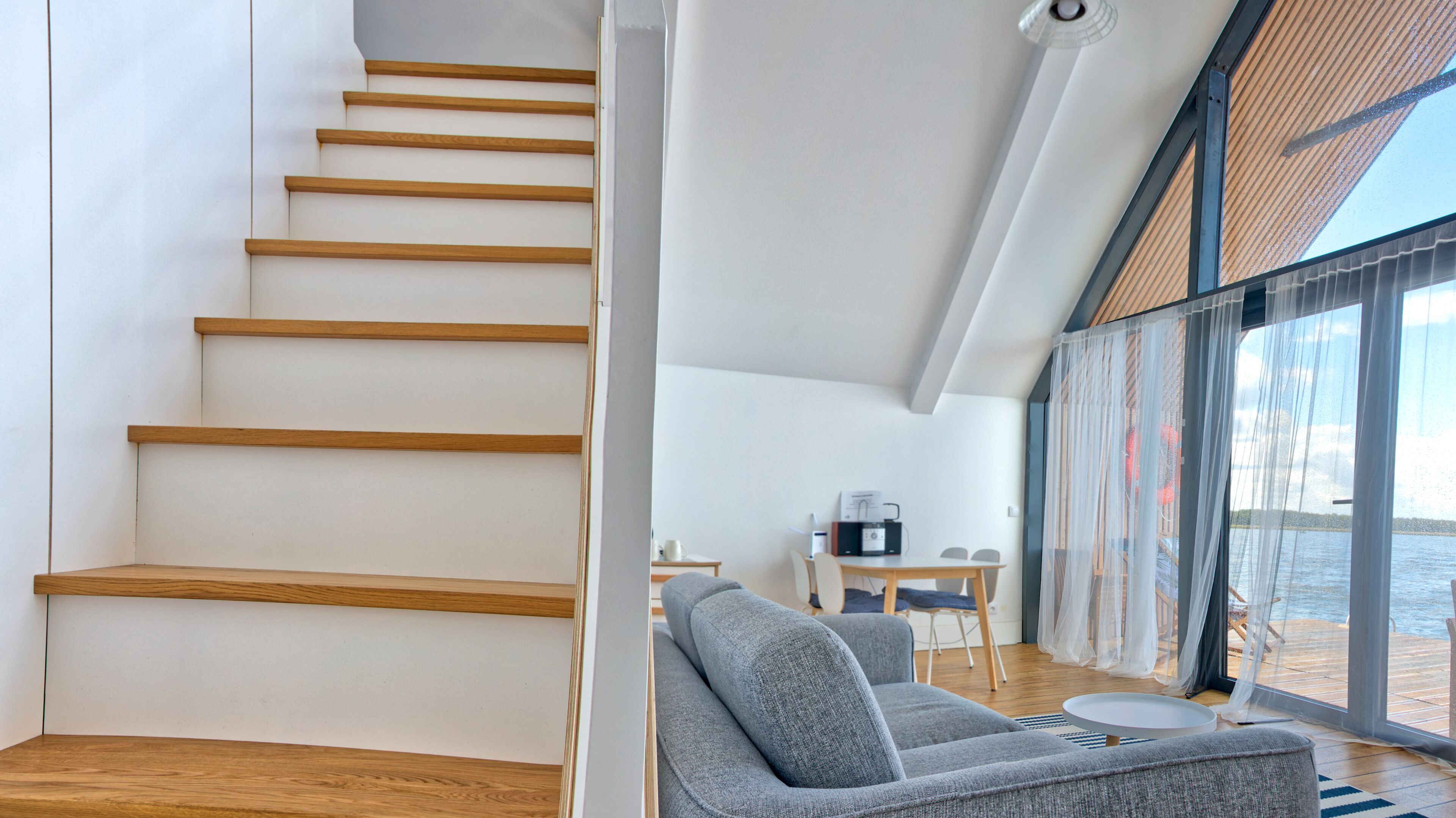 A modern interior with a wooden staircase, a gray sofa, and a view of the sea through large windows.