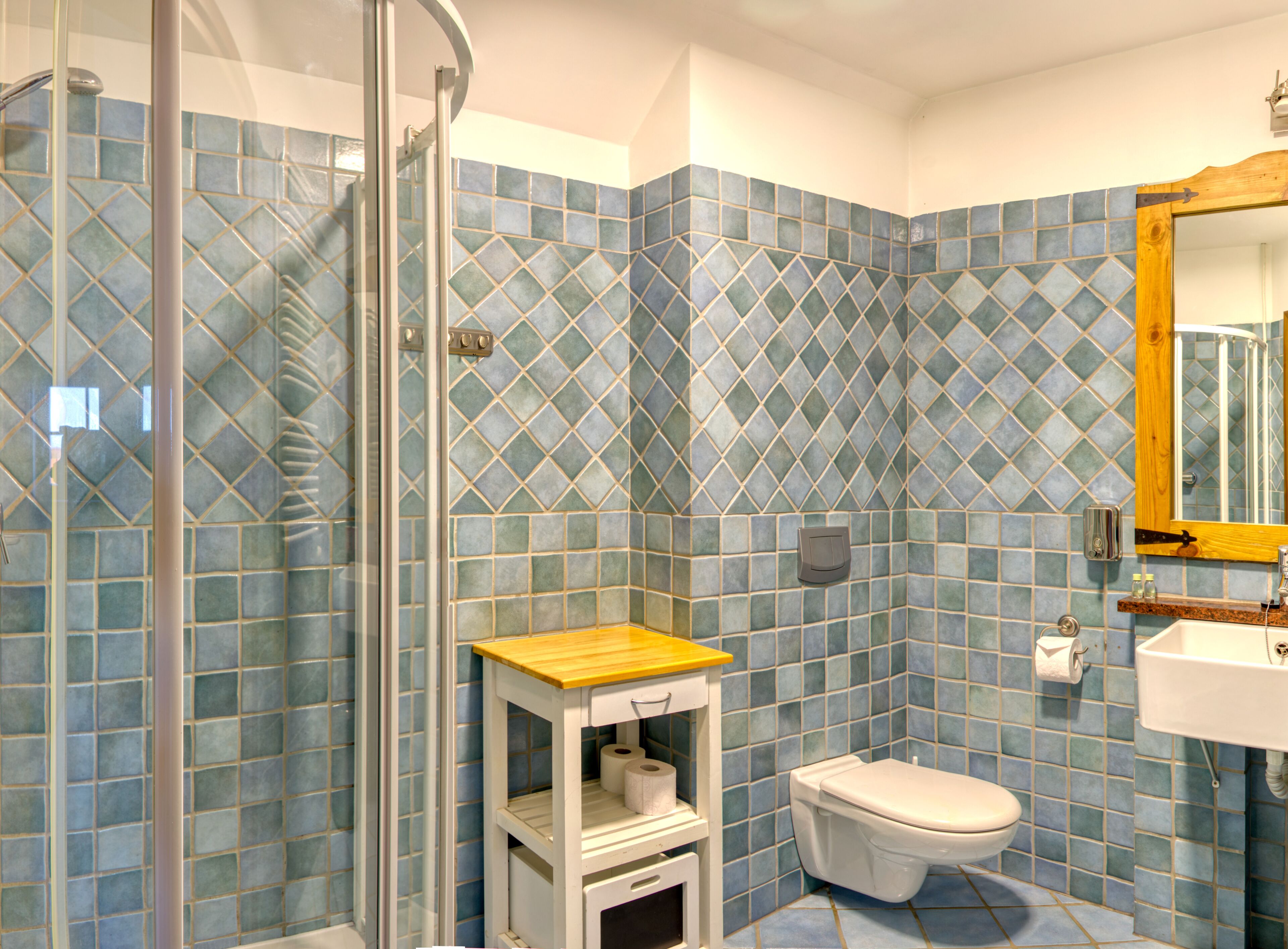 A bathroom with blue mosaic tiles, a glass shower enclosure, a wooden vanity, and a white toilet.