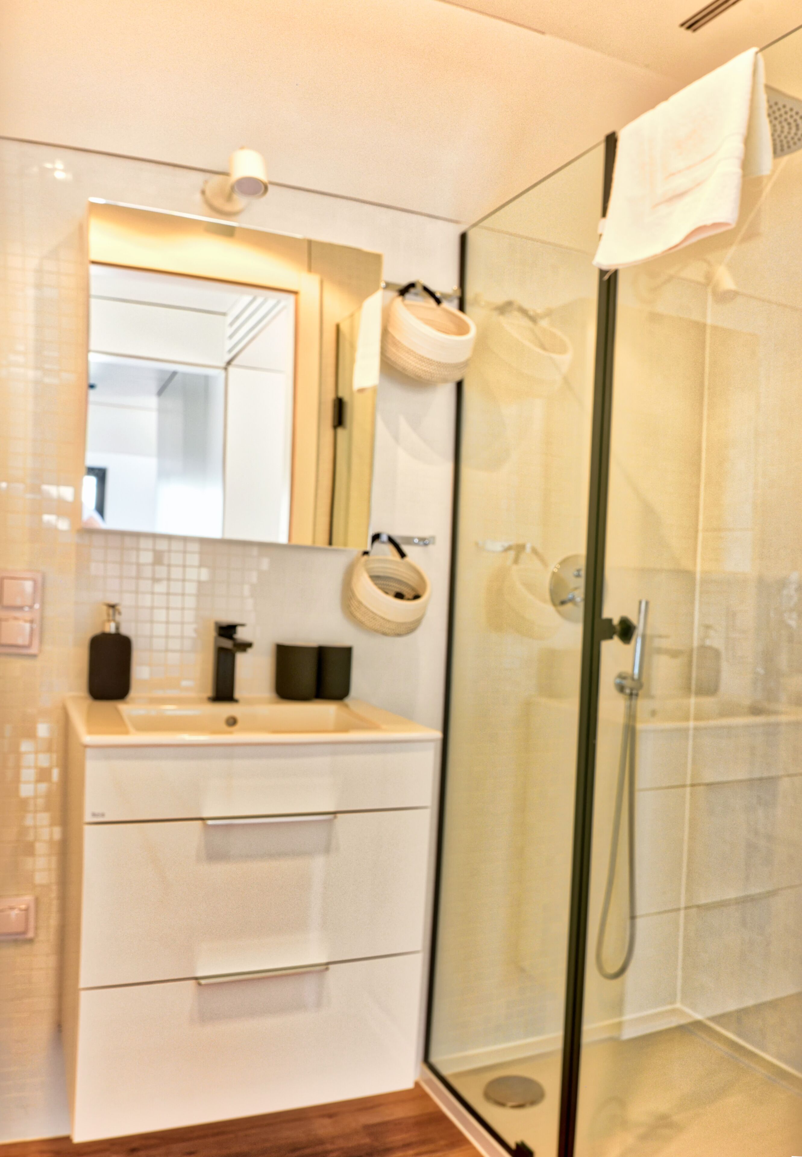 Un baño moderno con cabina de ducha de vidrio, lavamanos blanco con espejo y suelo de madera.