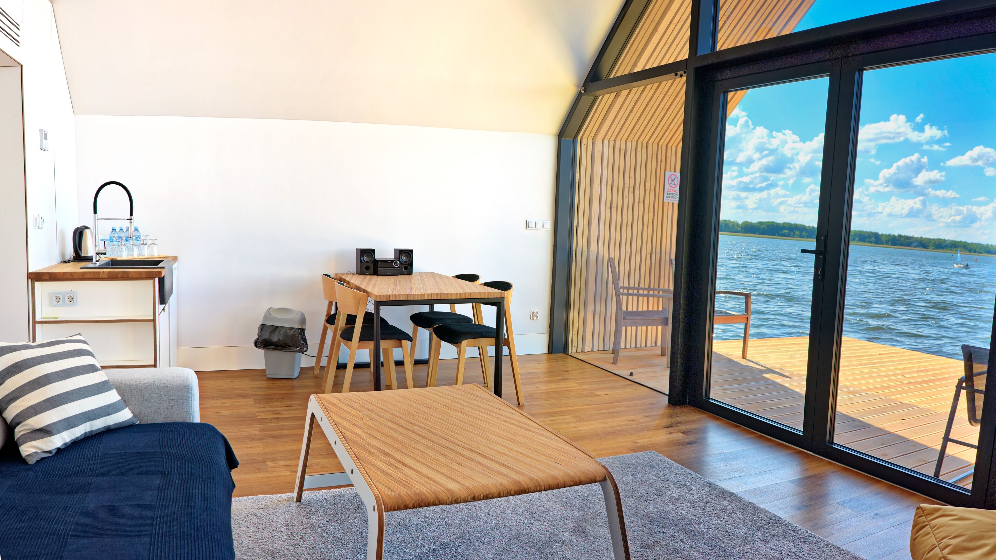 A modern room with a wooden table, chairs, a sofa, and a large window overlooking a lake.