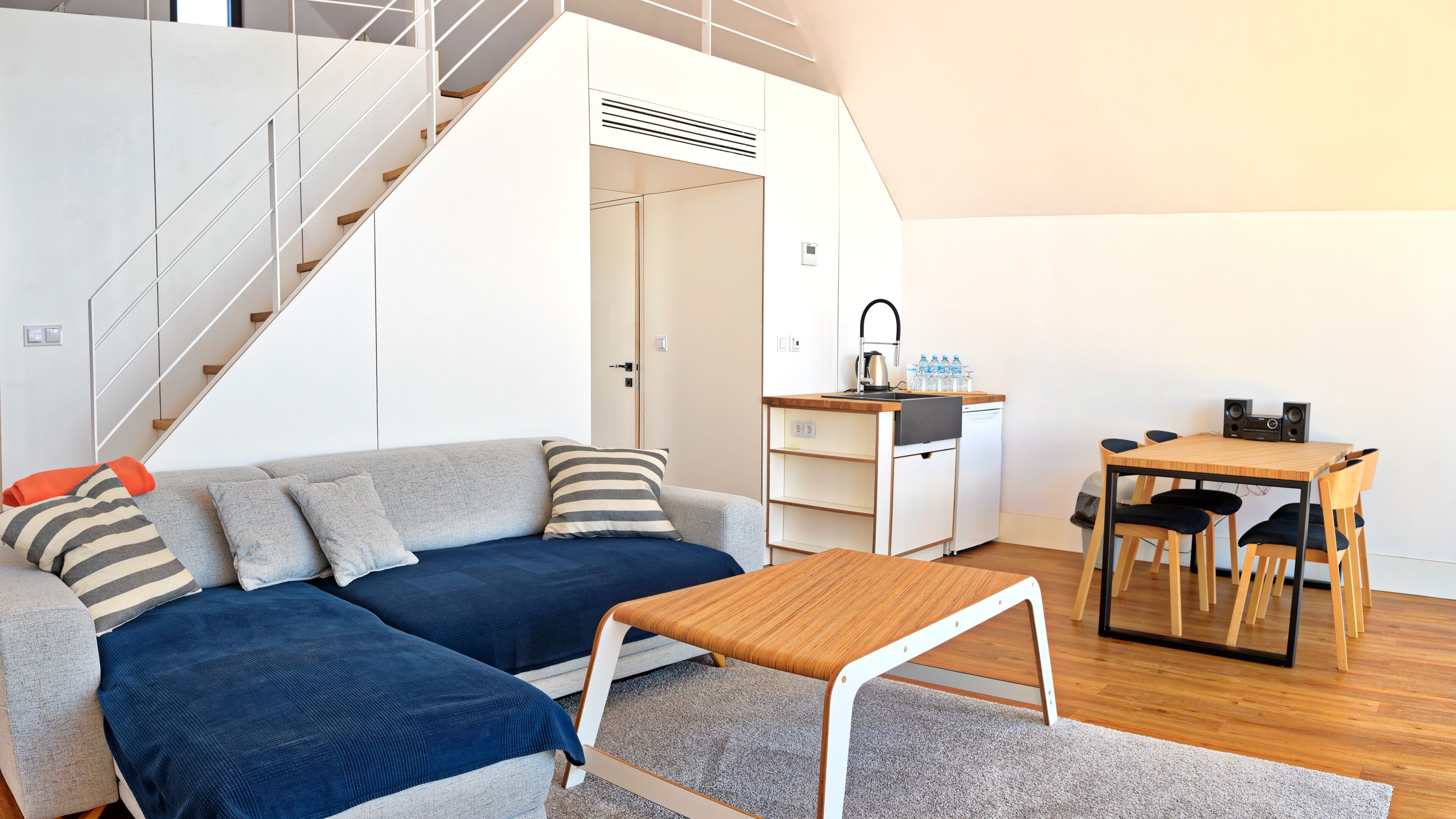 Un espacio habitable moderno con escalera, un conjunto de sofás, una mesa de comedor y una kitchenette.