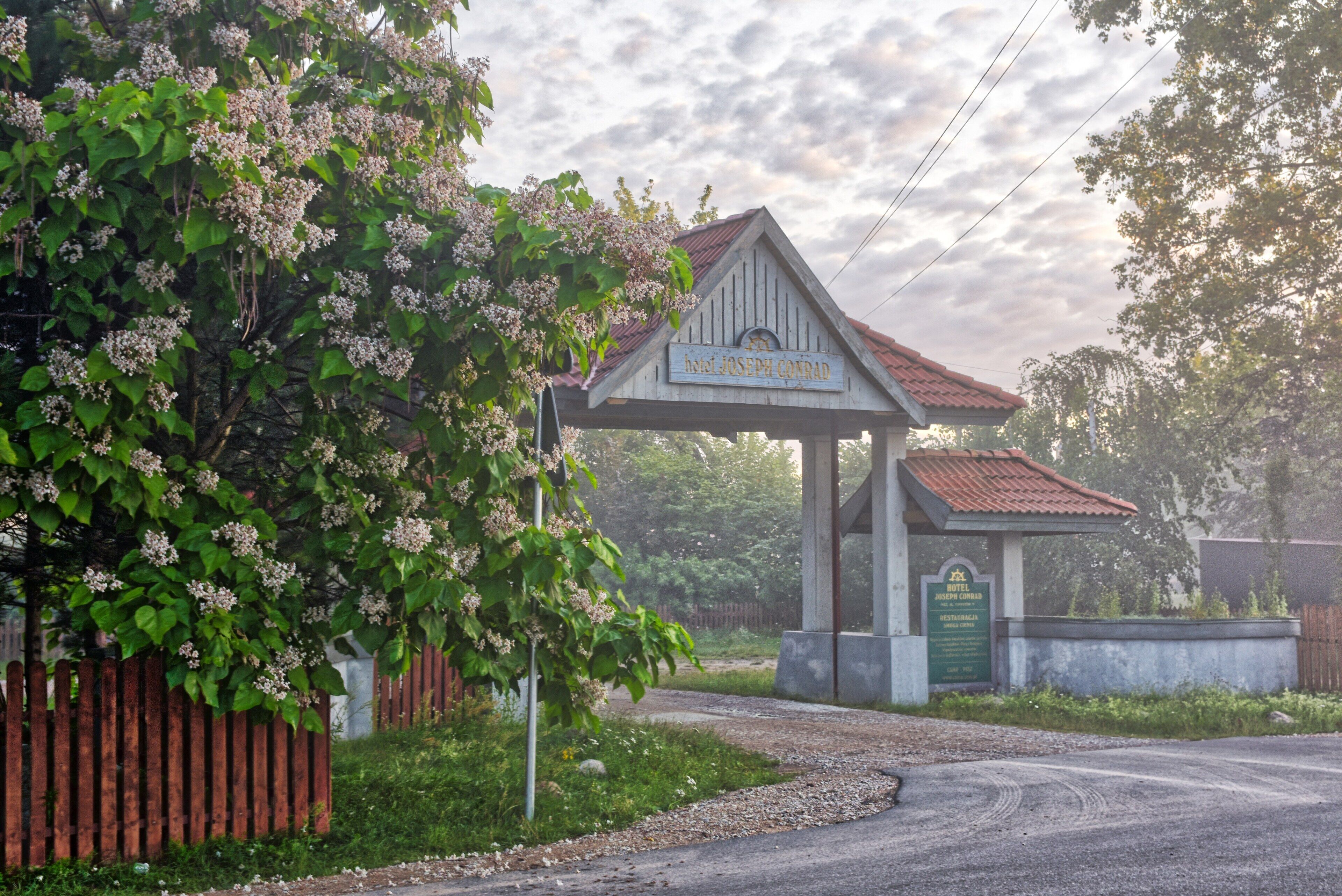 A hotel entrance with a sign reading "Hotel Joseph Conrad," surrounded by lush greenery and a wooden fence.