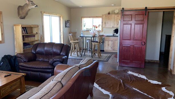 A spacious living room with a leather sofa, a dining area with wooden chairs, a kitchen with wooden cabinets, and a red door.