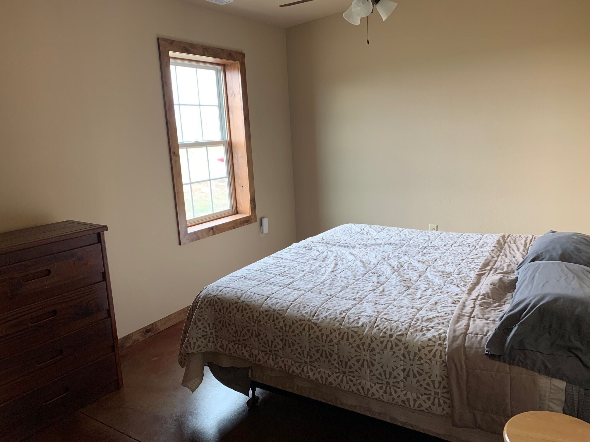 Une chambre à coucher avec un lit, une commode en bois, une fenêtre et un ventilateur de plafond.