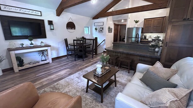 A spacious living room with a white sofa, a brown leather chair, and a wooden coffee table. A dining area with a wooden table and chairs is visible, along with a kitchen area featuring a stainless steel refrigerator and wooden cabinets. - Get away from the stress and strain of life at this romantic mountain cabin. (Ronan)