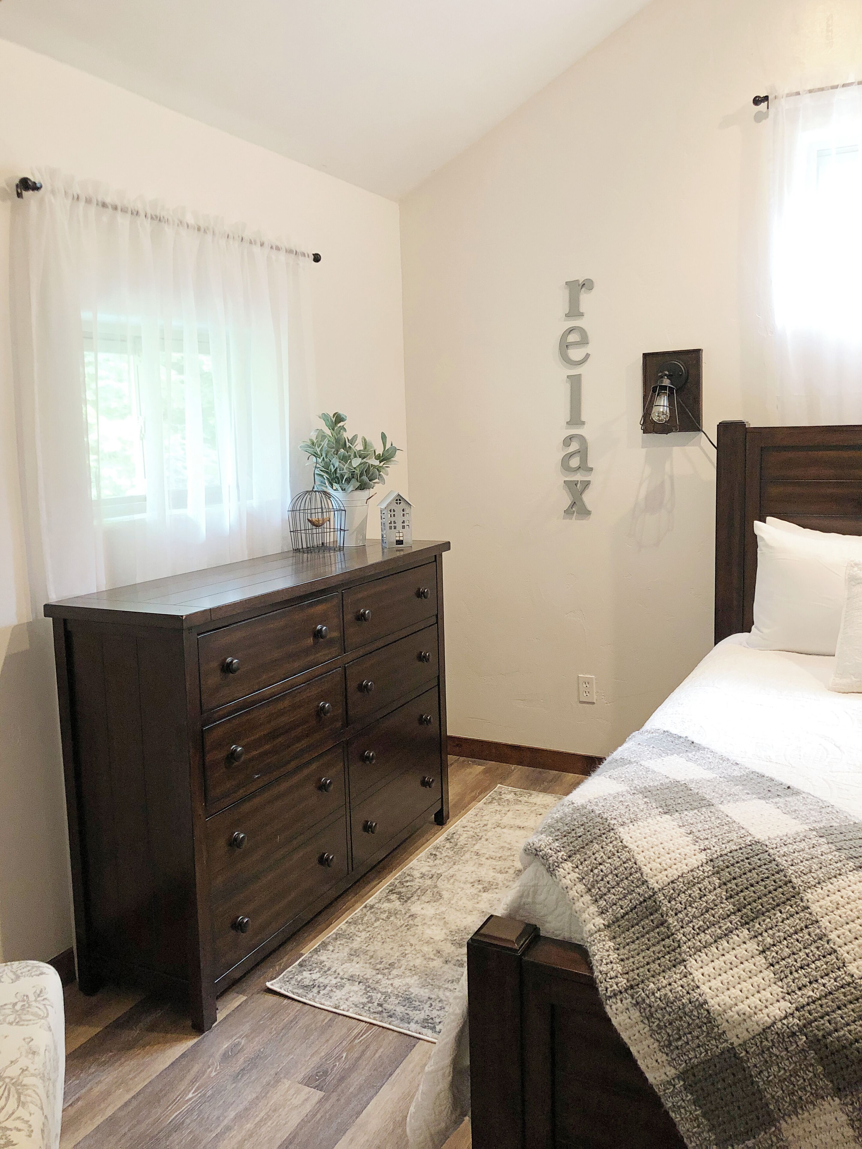 A bedroom with a wooden dresser, a bed with a checkered blanket, a wall-mounted light fixture, and a small potted plant.