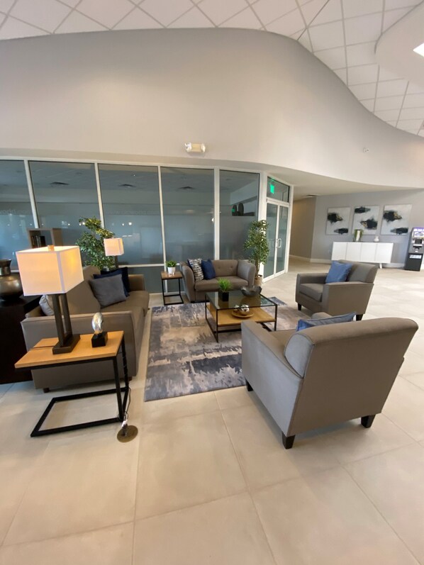 A modern lobby with a curved ceiling, seating area with sofas and armchairs, a coffee table, and a lamp. Large windows provide natural light, and there is a reception desk in the background.