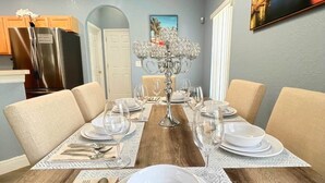 A dining room with a wooden table set for four, surrounded by beige chairs. A chandelier hangs above, and a painting of a cityscape is on the wall.