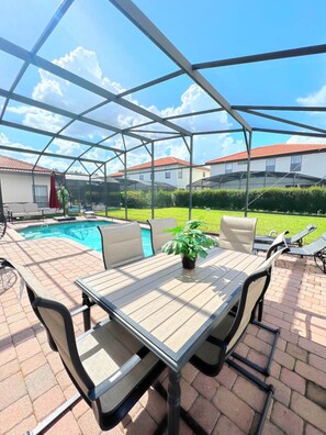 Um pátio externo com mesa e cadeiras cobertas, piscina e vista para casas ao fundo.