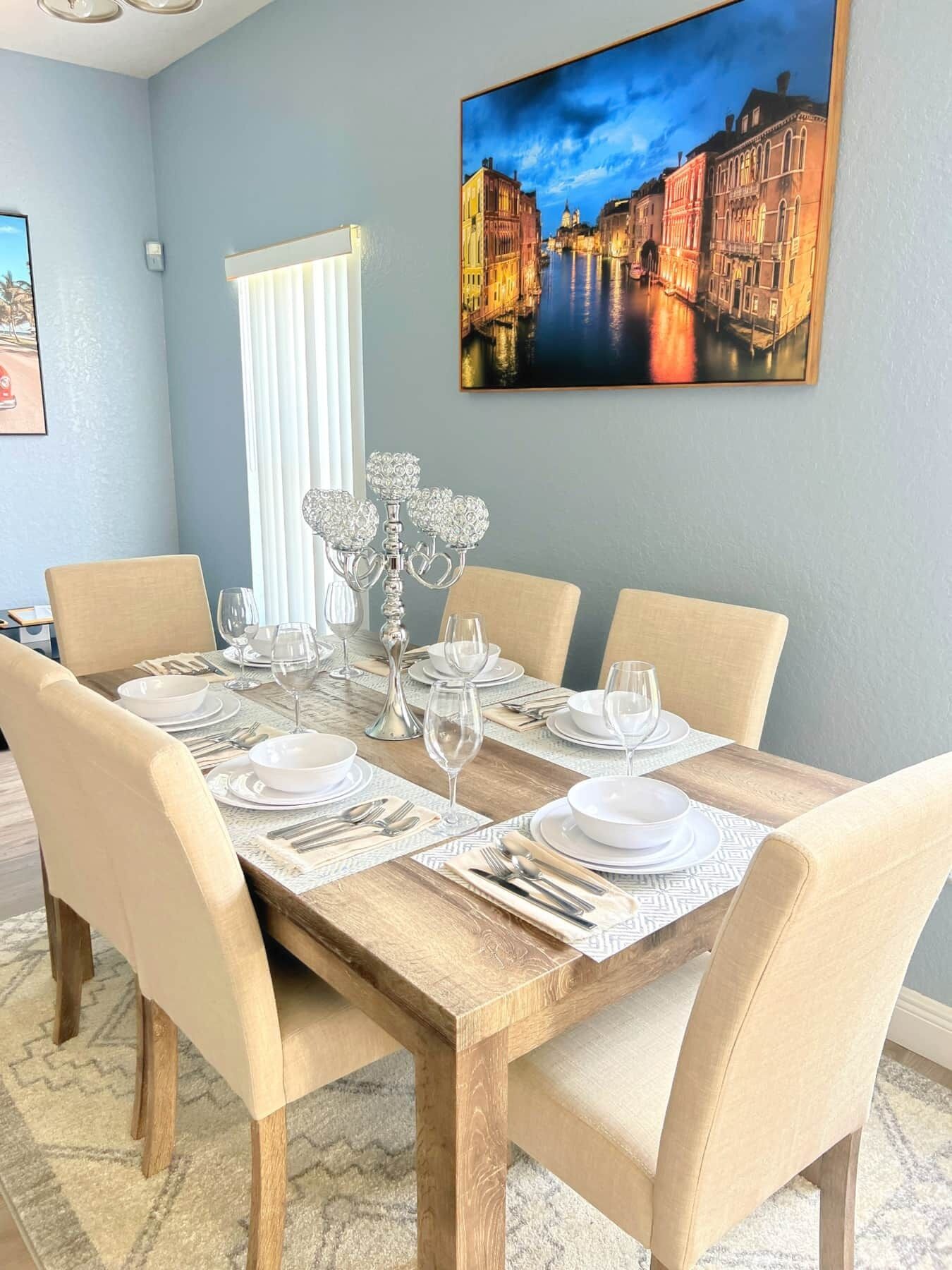 A dining room with a wooden table set for six, surrounded by beige chairs. A large painting of a cityscape is on the wall above the table.