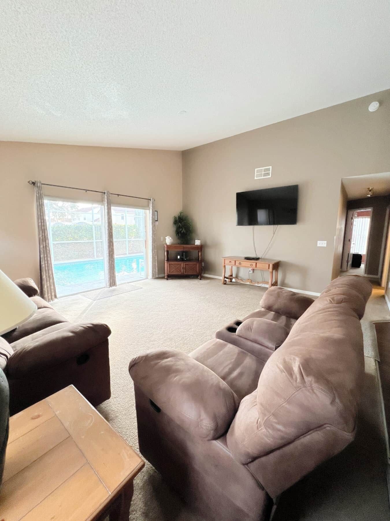 A living room with a large sectional sofa, a flat-screen TV mounted on the wall, and a sliding glass door leading to a balcony.