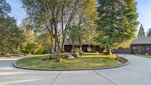 Un camino de entrada residencial con un césped circular, una casa de madera y un árbol grande.