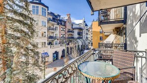 A balcony with wicker furniture and a glass table, overlooking a street with buildings and a tree. - Vail Mtn Views, 100m to Gondola | LH Studio (Vail)