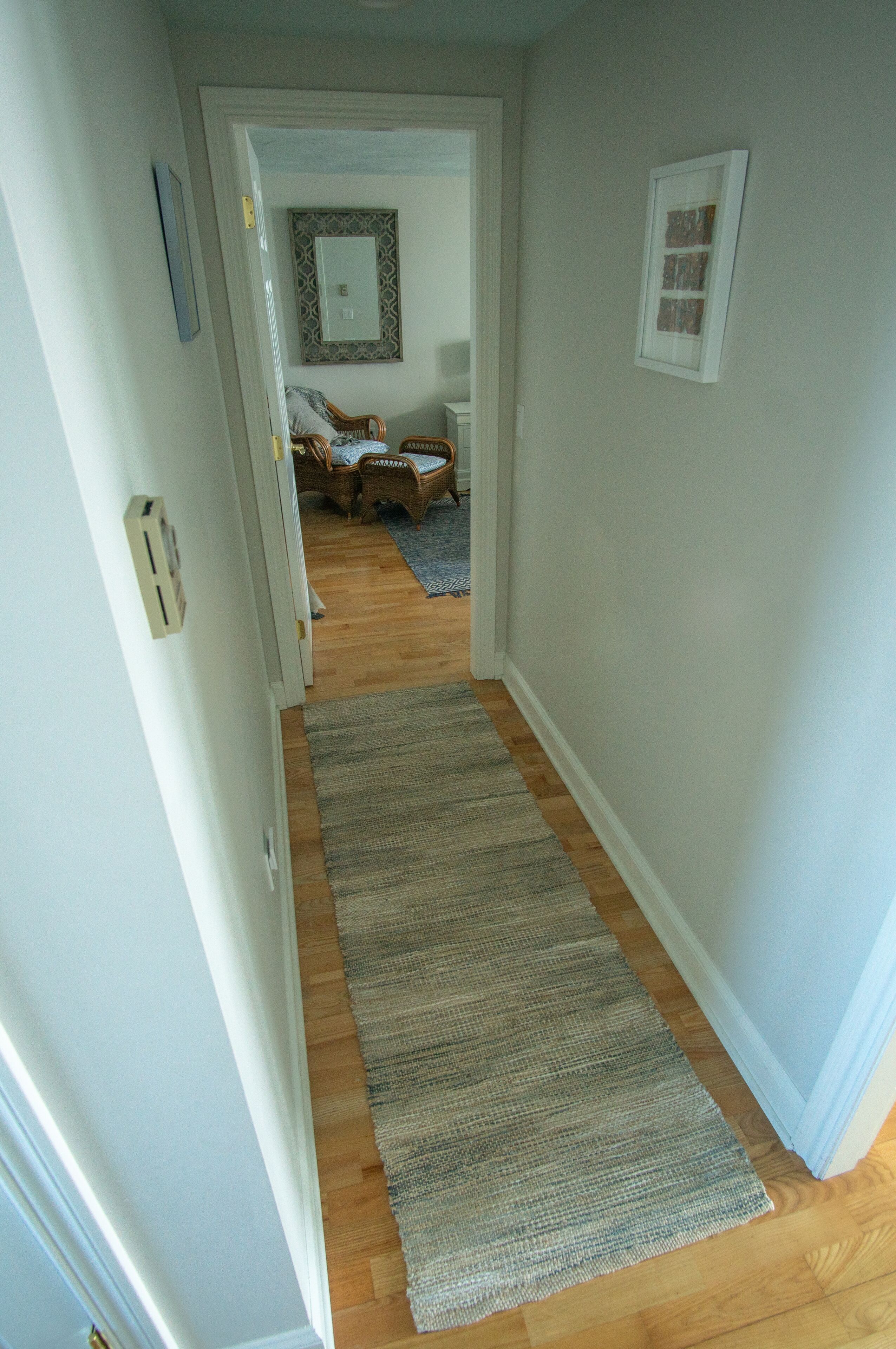 A hallway with a wooden floor, a gray rug, and a door leading to a room with a patterned rug and a wicker chair.
