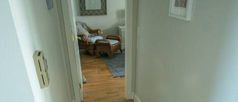 A hallway with a wooden floor, a gray rug, and a door leading to a room with a patterned rug and a wicker chair.
