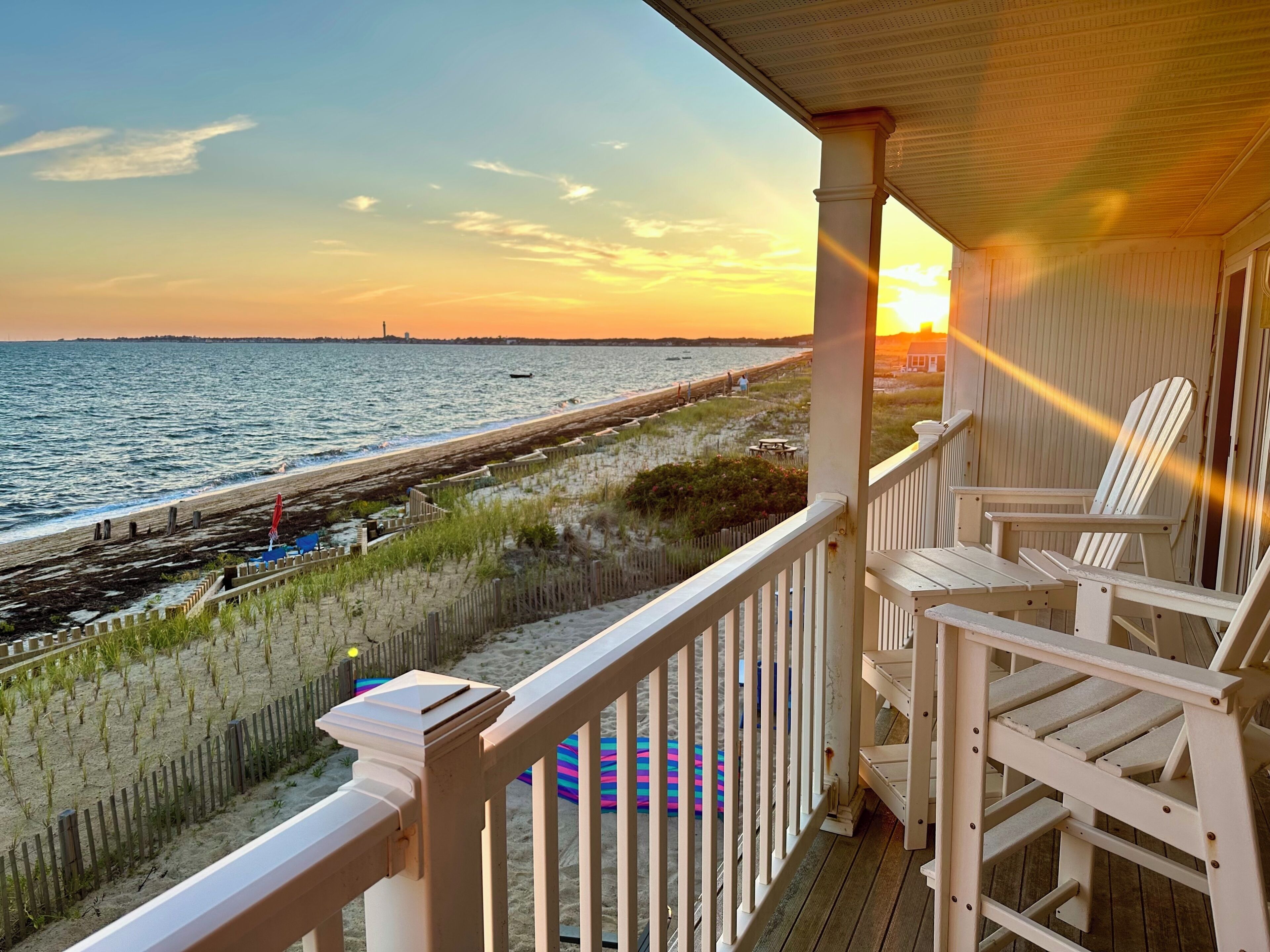 A wooden deck with white railings overlooking a beach with a fence and a pier.