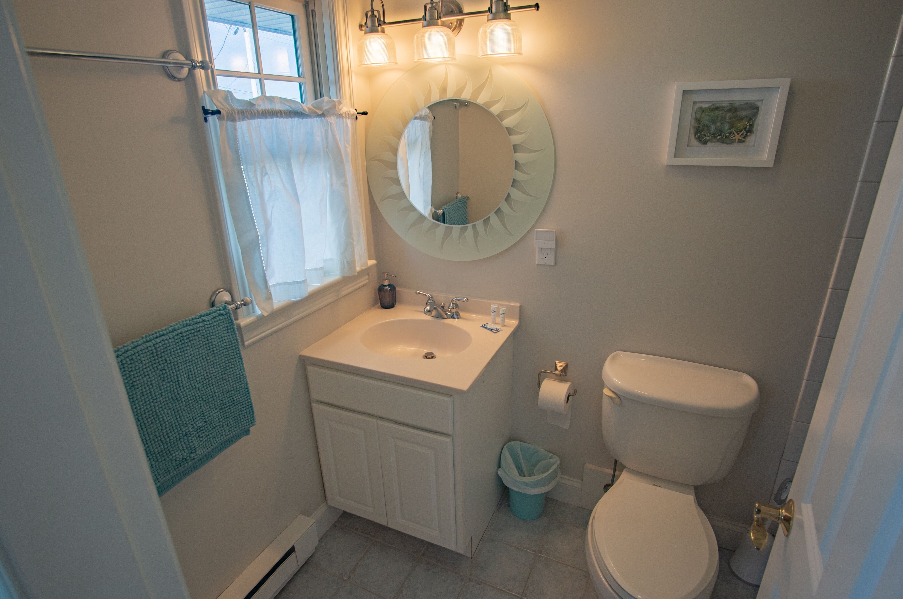 A bathroom with a sink, mirror, toilet, and a window with a curtain.