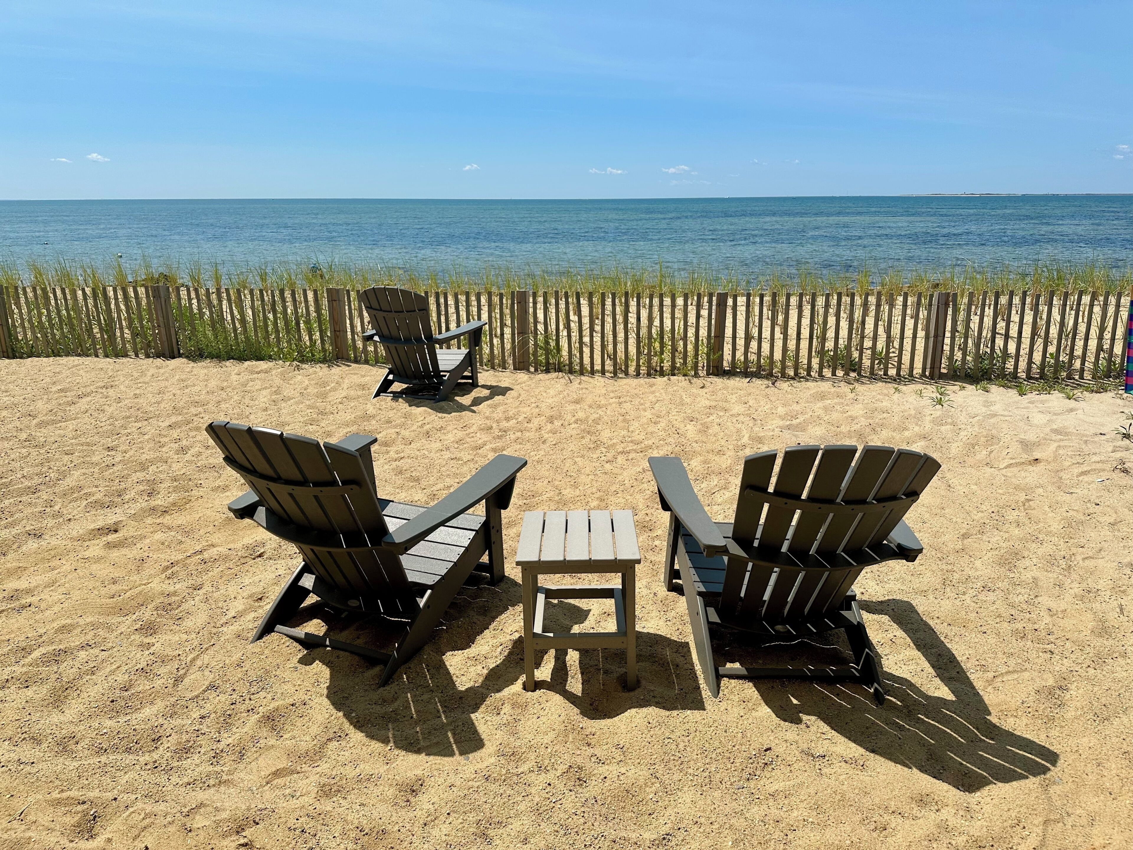 A beach setting with three Adirondack chairs and a small table, a wooden fence in the background, and a view of the ocean.