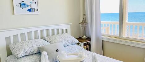 A bedroom with a bed, pillows, a bedside table with a lamp, a painting of fish, and a view of the sea through a window.