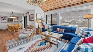 Un salón amplio con un sofá seccional azul grande, una mesa de comedor de madera y sillas. Un candelabro cuelga del techo y hay ventanas grandes con vistas a un paisaje nevado.