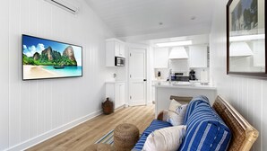 Una habitación moderna y bien iluminada, con un televisor de pantalla plana mostrando una escena de playa tropical, una kitchenette con armarios blancos, piso de madera y una zona de estar acogedora con un sofá estampado.