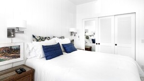 Dormitorio en tonos blancos con una cama grande, dos lámparas fijadas a la pared, un cuadro enmarcado y una mesita de mimbre con dos controles remotos.