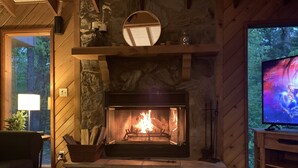 Un salon chaleureux avec une cheminée en pierre, un téléviseur et une fenêtre donnant sur une forêt.