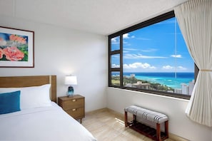 A hotel room with a bed, bedside table, bench, and a large window with a city and ocean view. - Waikiki Banyan w/ lanai & beach access (Honolulu)