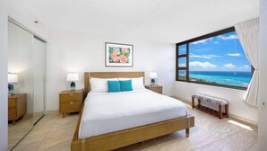 Une chambre avec un grand lit, des tables de chevet, un banc, une fenêtre donnant sur la mer et un tableau au mur.