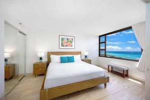 A bedroom with a large bed, bedside tables, a bench, a window with a view of the sea, and a painting on the wall.