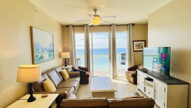 A hotel room with a sofa, a coffee table, a TV, and a balcony with a view of the ocean.