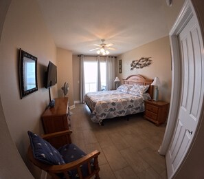 Quarto com cama, poltrona, penteadeira, mesa de cabeceira, luminária, ventilador de teto e porta.