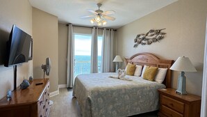 A hotel room with a bed, a TV, a wooden dresser, a desk, and a view of the ocean.