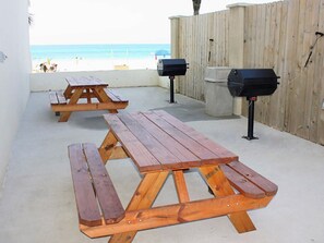 Pátio com mesas de piquenique e churrasqueiras, com vista para uma praia.
