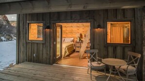 Una cabaña de madera con un porche de madera, un comedor con mesa y sillas, y un dormitorio con cama y cómoda.