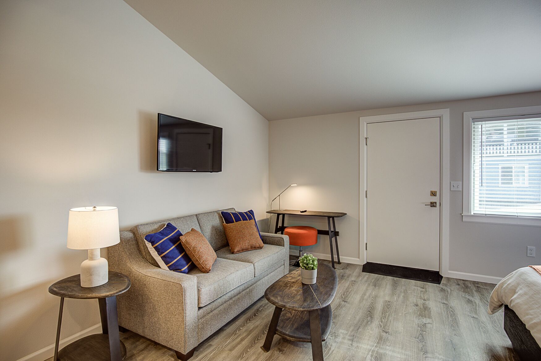 A living room with a sofa, two side tables, a coffee table, a TV mounted on the wall, a door, and a window with blinds.