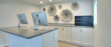Uma cozinha moderna com dois iMacs da Apple em uma bancada, armários brancos e arte decorativa na parede.