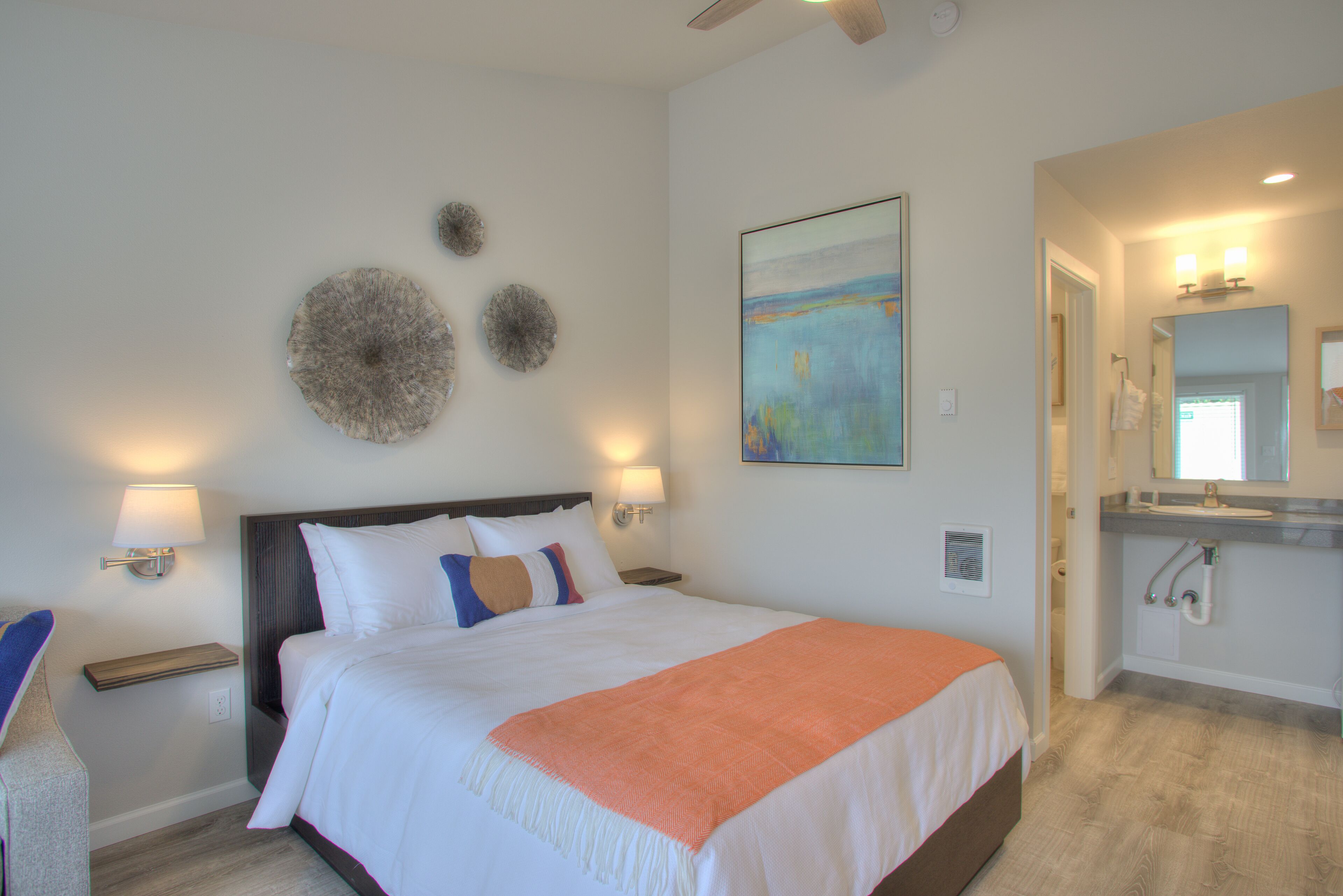 A hotel room with a large bed, two bedside lamps, a wall-mounted artwork, and a bathroom with a sink and mirror.