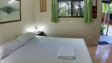 A single bed with white linens, a nightstand with a lamp, a framed picture of a giraffe, a window with green curtains, and a view of trees and a bridge outside.