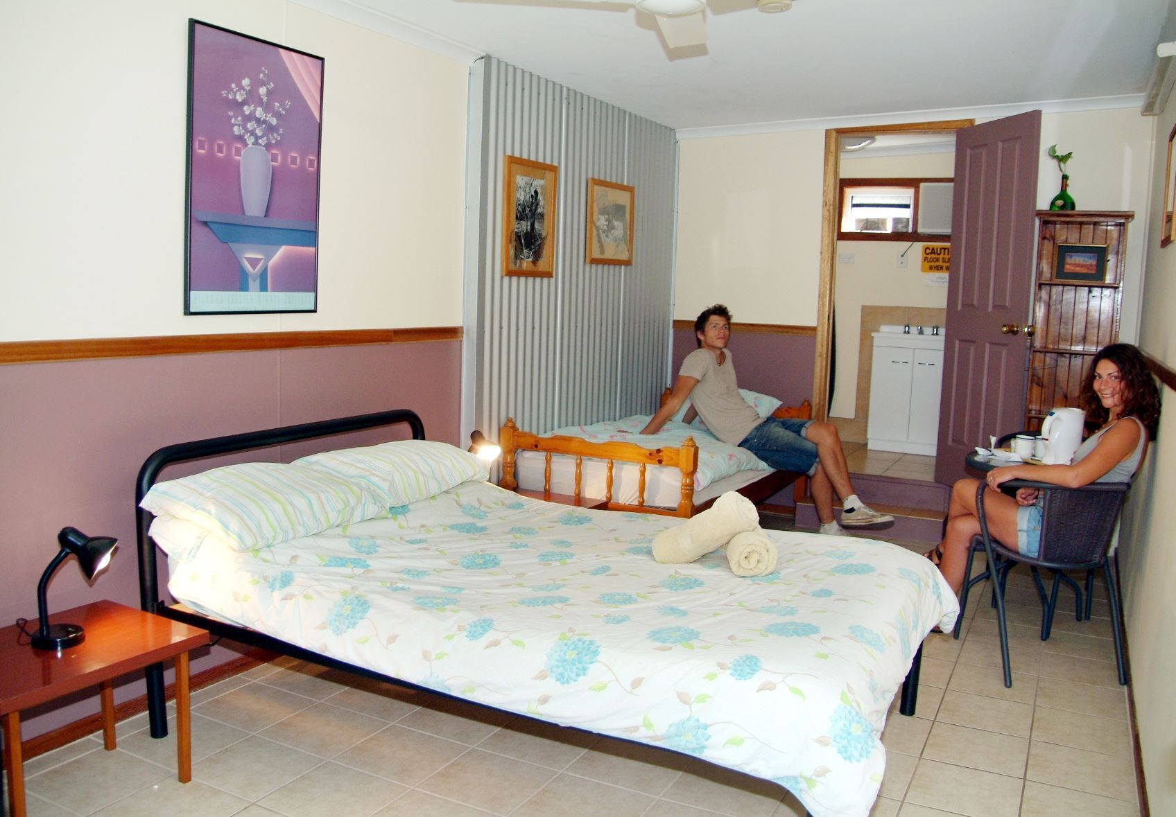 A hotel room with a large bed, a bedside table, a chair, and a small kitchenette area.