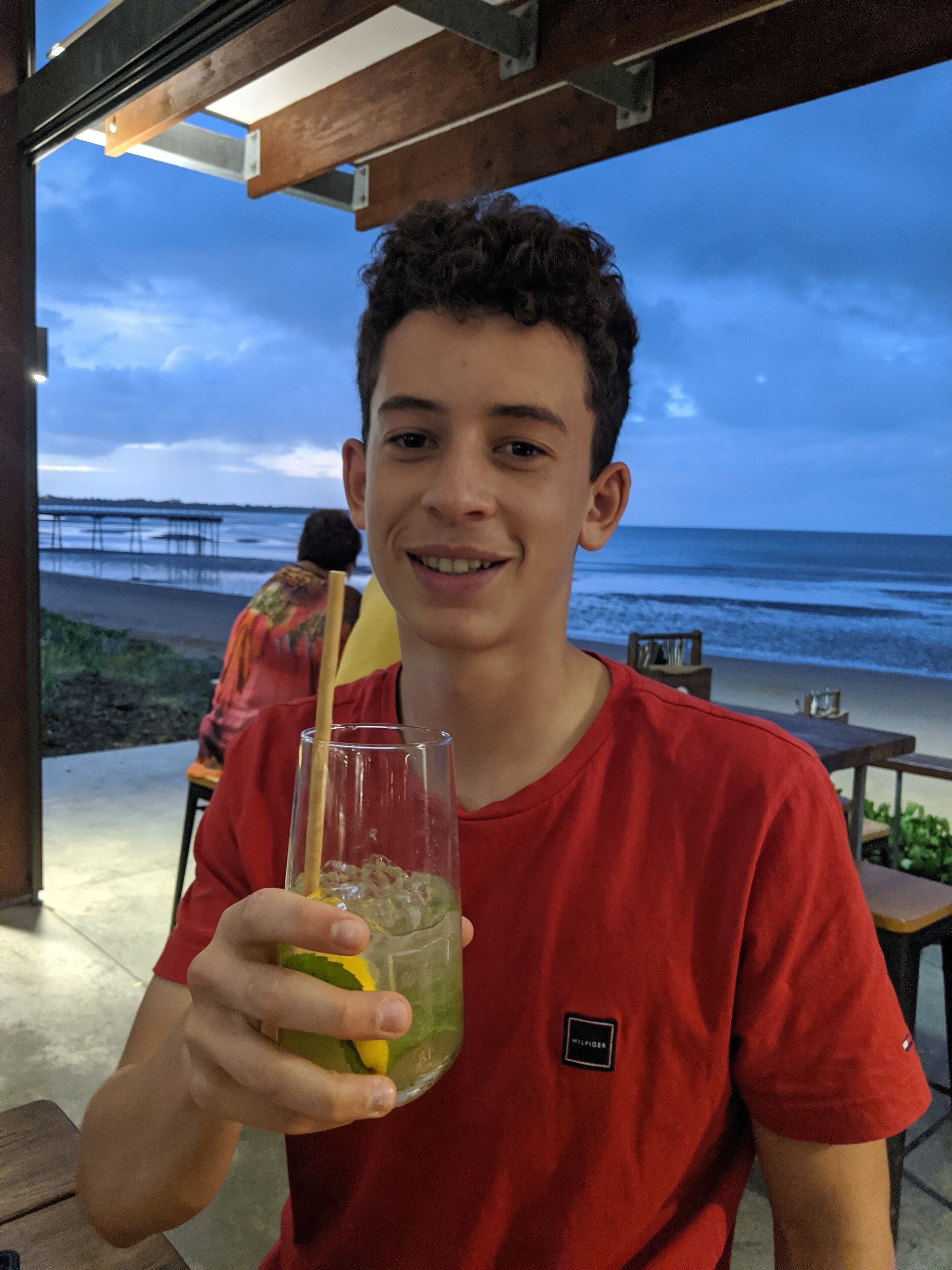 Une personne tenant un verre muni d’une paille, vêtue d’un t-shirt rouge, assise à une table en plein air, avec la plage et l’océan en arrière-plan.