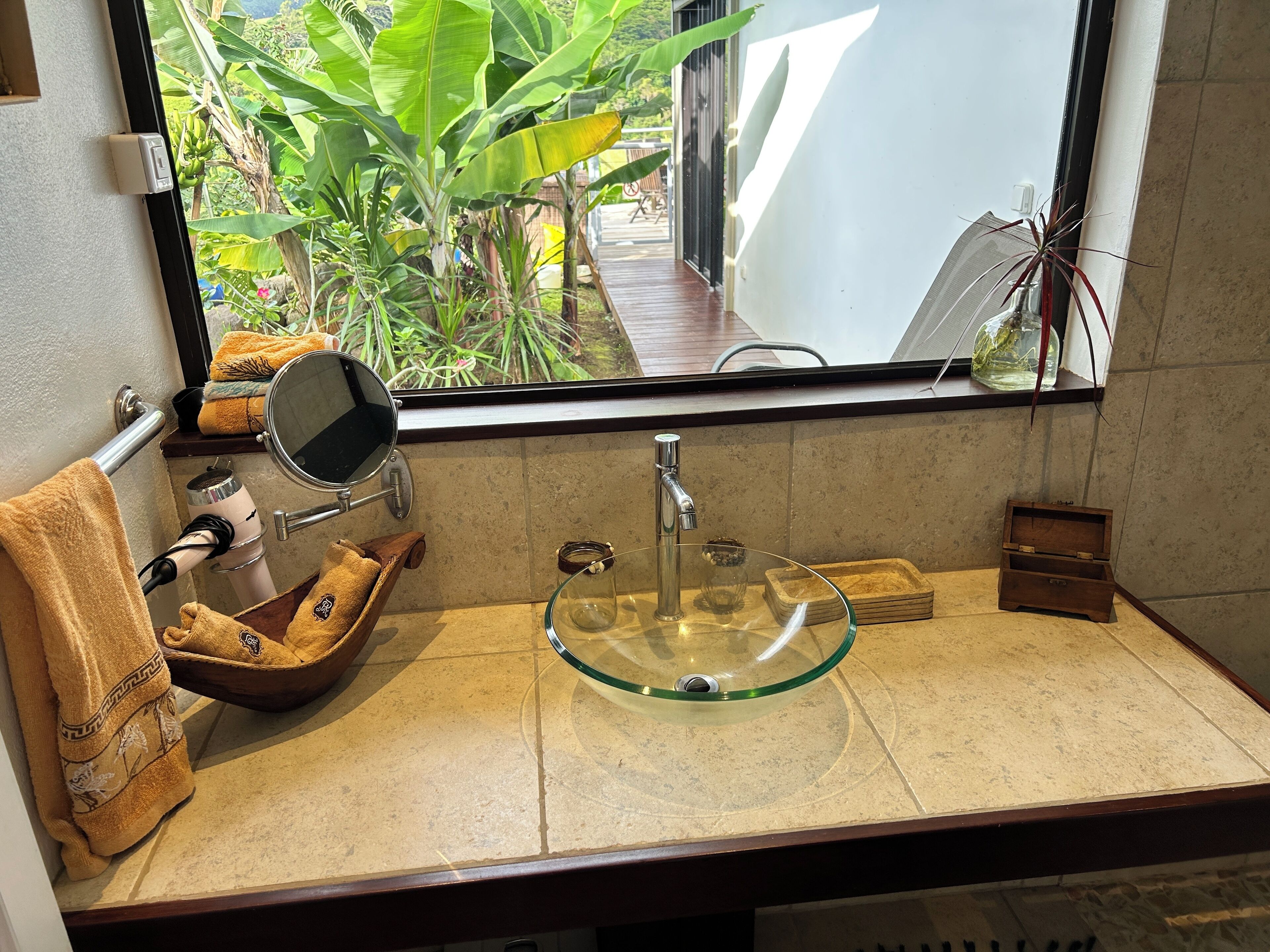 Une salle de bain avec un lavabo en verre, un porte-serviettes sur lequel est posée une serviette marron, un miroir et une fenêtre donnant sur un paysage verdoyant.