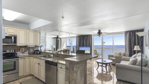 A modern kitchen with granite countertops, stainless steel appliances, and a large island. There is a dining area with a round table and chairs, and a living room with a sofa and a TV. The room has large windows with a view of the ocean. - 18th Floor Pelican Beachfront with Ocean View (Destin)