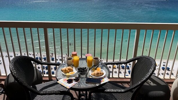 A balcony with a table set for breakfast, overlooking a beach with people and a clear view of the ocean. - 18th Floor Pelican Beachfront with Ocean View (Destin)