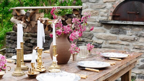 A rustic outdoor dining setup with a stone fireplace, wooden table, and candle holders.